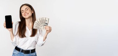 Portrait of smiling woman showing mobile phone screen and money cash, looking happy, standing over white background.