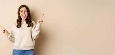 Cheerful caucasian woman showing directions, two ways, pointing sideways at variants, choices for customer, standing over beige background.