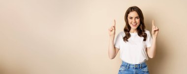 Enthusiastic young woman, female customer pointing fingers up and smiling, showing banner or logo, standing against beige background.