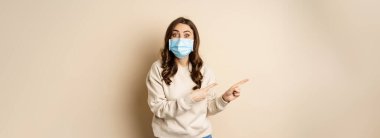 Health and covid-19 concept. Excited woman in medical face mask, pointing fingers left and looking amazed, reacting impressed with promo offer, beige background.