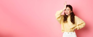 Sinirli ve hayal kırıklığına uğramış genç bir kadın yüzü avuçlarında, alnının üzerinde el ele tutuşuyor ve hayal kırıklığına uğramış bir şekilde kameraya bakıyor, pembe arka planda duruyor..