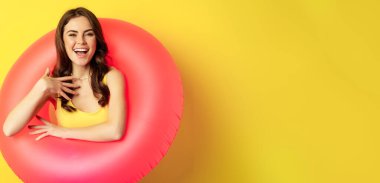 Close up portrait of beautiful brunette woman model inside pink swimming ring, beach accessories and summer vacation concept, yellow background.