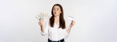 Excited woman holding credit card and money, looking amazed at cash, standing against white background. Copy space