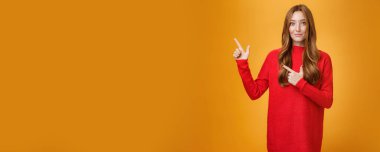 Yumuşak ve nazik, kızıl saçlı, kırmızı, sıcak kazaklı, mutlu bir şekilde gülümseyerek sol üst köşeyi gösteren, ilginç bir yer gösteren, turuncu arka plan üzerinden ziyaret etmeyi öneren kız..