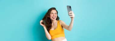 Teknoloji ve yaşam tarzı konsepti. Mutlu genç bir kadın akıllı telefon uygulamasında filtreli selfie çekerken aptal suratlar yapıyor, mavi arka planda duruyor..