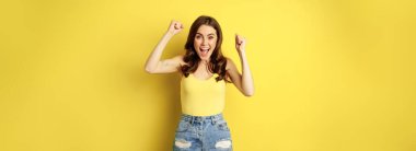 Enthusiastic young woman shouting and cheering, rejoicing, raising hands up and screaming in joy, chanting, standing over yellow background.