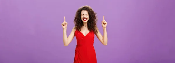 Life changing moment happening now. Joyful and happy overwhelmed good-looking elegant woman with curly hairstyle in red evening dress closing eyes grinning from happiness, pointing up over purple wall