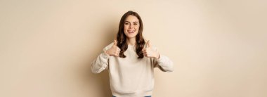 Beautiful young woman, student showing thumbs up in approval, recommending store, standing over beige background.
