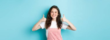 Neşeli esmer kızın portresi, başparmağını kaldırıyor, gülümsüyor ve iyi işi övüyor, ürünü onaylıyor ve tavsiye ediyor, mavi arka planda duruyor..