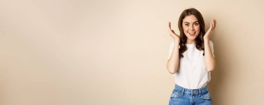 Surprise. Happy young woman open eyes with hands and waiting for gift, standing over beige background. Copy space