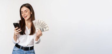 Portrait of happy smiling woman using mobile phone app, holding money cash, standing over white background. Copy space