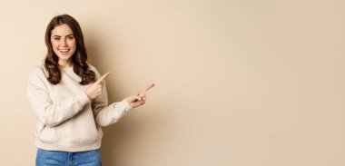 Smiling gorgeous woman pointing fingers right, inviting people, showing logo or banner, standing over beige background.