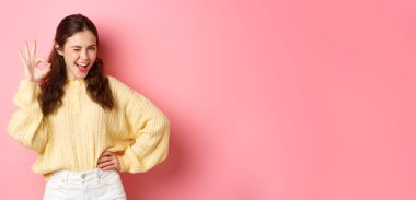 Küstah genç kadın göz kırpıyor, işaret veriyor, onaylıyor ve onaylıyor. Kız izin vermek için iyi bir hareket yapıyor, evet diyor, pembe arka planda duruyor..