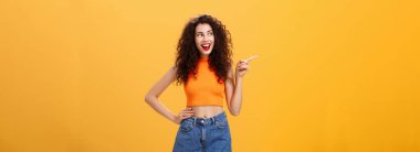 Coşkulu, stil sahibi genç bir kadının portresi. Kırmızı rujlu kıvırcık saç stili ve bel üstü açık el ele tutuşarak turuncu arka planda neşeli ve mutlu görünüyor..