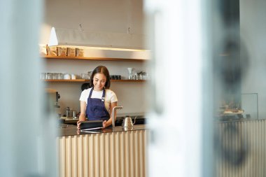 Kahve dükkanı. Gülümseyen Koreli barista, tezgahın arkasında çalışan kadın, kafede sipariş almak için tablet kullanıyor, iş yerinde mavi önlük giyiyor..