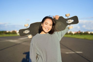 Boş zaman ve insanlar. Asyalı mutlu bir kadın, elinde uzun panoyla kırsalda boş bir yolda geziyor. Kaykaycı kız kaykayını tutuyor ve kameraya gülümsüyor..
