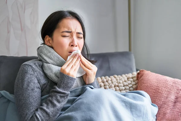Image of sick korean woman at home, covered in warm clothes and scarf, feeling sick, catching a cold and sitting on sofa sneezing.