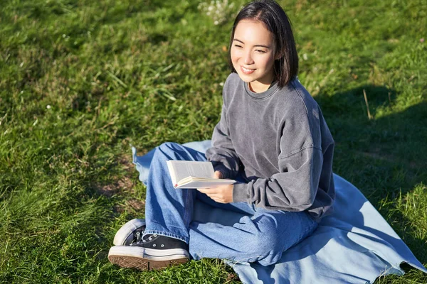 Portrait of asian girl reading book, sitting on her blanket in park, with green grass, smiling happily.