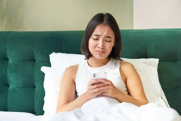 Disappointed asian girl, crying and whining, feeling upset, lying in bed on pillow, holding mobile phone, read upsetting bad news, waking up in awful mood.