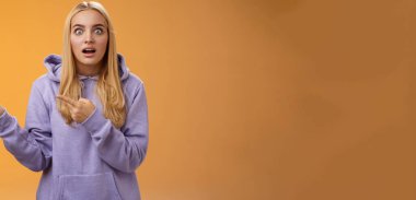 Kapüşonlu, sarışın, şaşkın bakışlı, şaşırmış, şaşırmış bakışlı, ağzı açık, solu işaret eden, dedikoducu, turuncu arka planda duran sarışın kadın şaşırmış..