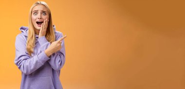 Şaşırtıcı derecede heyecanlı kapüşonlu genç sarışın kız avucunun içi gibi bağırıyor sağa doğru hayret verici bir şekilde haykırıyor ünlü süper star neşeli, pozitif duyguları tutamıyor, turuncu arka plan.