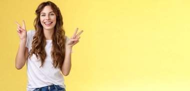 Dost canlısı, sevimli, mutlu, kıvırcık, uzun kıvırcık saçlı, barış zafer işaretleri gösteren, hareketli poz veren, neşeli, kameralı, gülen, sarı arka planda duran genç bir kız. Yaşam biçimi.