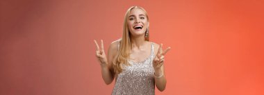 Neşeli, çekici, beyaz, beyaz kadın model, gümüş renkli, gösterişli elbiseler içinde. Barış zaferi jesti, gülümsüyor, doğum günü partisinde eğleniyor, kırmızı arka planda duruyor..