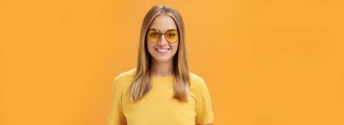 Zarif, kendine güvenen, sarışın, bronz tenli, sarı tişörtlü, güneş gözlüklü, neşeli, turuncu arka planda poz veren ve kameraya gülümseyen Avrupalı bir kadın.