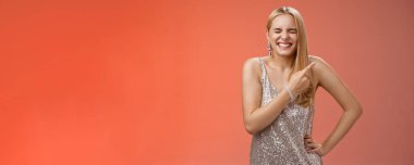 Kaygısız, neşeli, sarışın, çekici bir kadın eğleniyor, gülüyor, gülüyor, erkek arkadaş kıkırdıyor, kıkırdıyor, kıkırdıyor, gülümsüyor, sağ üst köşeyi işaret ediyor kırmızı arka planın arkasında..