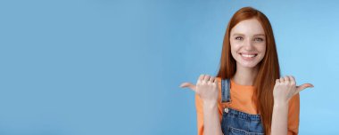 Kapalı alanda çekilen karizmatik iddialı güler yüzlü kızıl saçlı kadın turuncu gömlekli kot pantolon yan başparmakları sağa doğru işaret ediyor fırsatlar sunuyor seçenekler sunuyor şans, mavi arka plan.