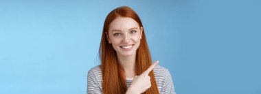 Yakın plan çekici, nazik, dost canlısı, güler yüzlü kızıl saçlı kadın sol üst köşedeki işaret parmağını göstererek sırıtarak mağaza bulmada yardımcı oluyor, mavi arka plan..