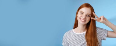 Neşeli, dost canlısı, kızıl saçlı, mutlu bakışlı, barış zafer işareti gösteren, başı eğik, sırıtan, beyaz dişli, neşeli, iyimser, mavi arka planda duran güzel kız..