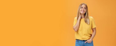 Sevimli ve sevimli Avrupalı bağımsız kadın, sarı tişörtlü, boynuna dokunarak iç çekiyor ve sağ üst köşede güzel bir gülümsemeyle turuncu arka planda poz veriyor..