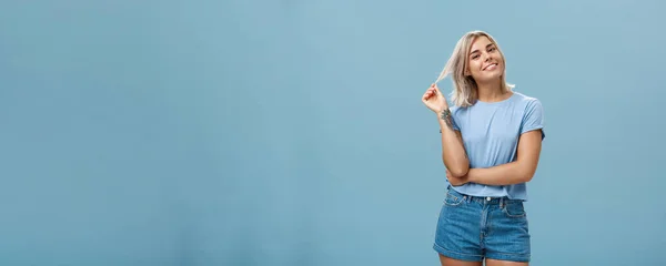 Studio shot of relaxed and chill popular attractive woman with blond hair and tattoos playing with strand tilting head and smiling as if listening boyfriend or flirting standing amused over blue wall