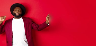 Noel ve insanlar kavramı. Mutlu siyah adam yeni yılı kutluyor ve dans ediyor, parti kıyafeti giyiyor, kırmızı arka plan.