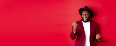 Şanslı Afro-Amerikan adam zaferi kutluyor, yumruklarıyla kazanıyor ve seviniyor, mutlu çığlıklar atıyor, gözleri kapalı, kırmızı arka plan.