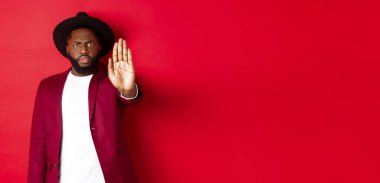 Ciddi ve endişeli siyahi adam seni durdurmak için elini uzatıyor, yasaklıyor ve karşı çıkıyor, somurtuyor ve surat asıyor, kırmızı arka planda dikiliyor..