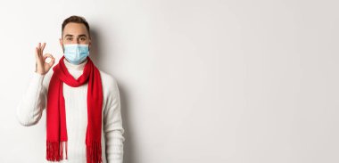 Covid-19, tecrit ve karantina konsepti. Yüz maskesi takmış tatmin olmuş yakışıklı adam işaret veriyor, onaylıyor ya da katılıyor, beyaz arka plan gibi..