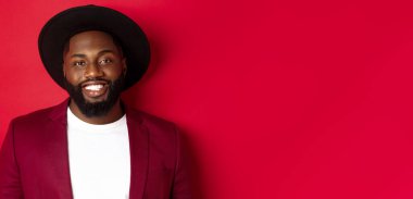 Sakallı, parti ceketi ve şık şapkalı, kameraya gülümseyen, kırmızı arka planlı yakışıklı Afro-Amerikan adam..