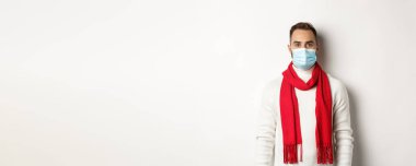 Covid-19, tecrit ve karantina konsepti. Beyaz arka planda duran, kışın giydiği süveter ve sağlık maskeli genç adam..