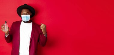 Covid-19, karantina ve tatil konsepti. Neşeli siyah adam kazanmaktan, hedefe ulaşmaktan, antiseptik göstermekten, tıbbi maske takmaktan, kırmızı arka planda durmaktan memnun..