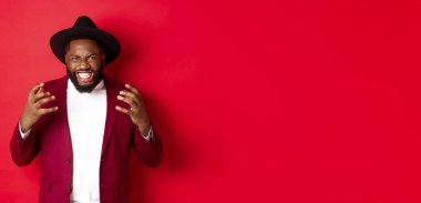 Kızgın Afrikalı Amerikalı adam agresif görünüyor, el sıkışıyor ve kameraya nefretle bakıyor, kırmızı arka plan.