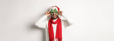 Noel, yeni yıl ve kutlama konsepti. Mutlu adam gülüyor, Noel Baba şapkası ve parti gözlüğü takıyor, beyaz arka planda duruyor..