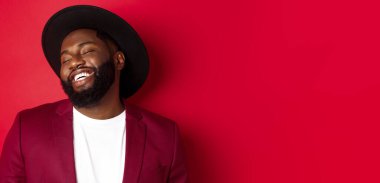 Yakından bakınca, uzun sakallı yakışıklı zenci adam gülüyor, eğleniyor, kaygısız görünüyor, kırmızı arka planda duruyor..