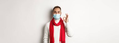 Covid-19, tecrit ve karantina konsepti. Yüz maskesi takmış tatmin olmuş yakışıklı adam işaret veriyor, onaylıyor ya da katılıyor, beyaz arka plan gibi..