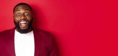 Afrika kökenli, sakallı, kameraya hayran kalmış, gülümseyen ve heyecanlı görünen, ceketle kırmızı arka planda duran Amerikalı adamın yakın çekimi..