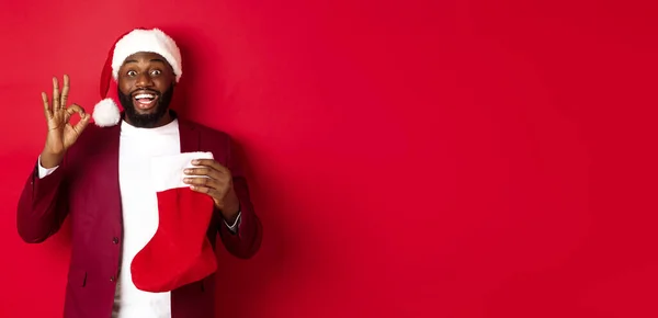 Amazed Black man holding holiday presents inside christmas sock, showing ok sign in approval, like something, standing over red background.