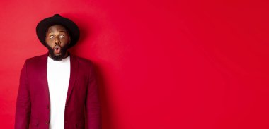 Moda ve parti konsepti. Şık giyinmiş etkileyici siyahi adam kameraya tamamen güvensiz bakıyor, soluk soluğa ve şaşırmış bakıyor, kırmızı arka planda duruyor..