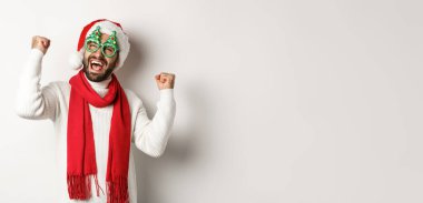 Noel, yeni yıl ve kutlama konsepti. Parti gözlüklü ve Noel Baba şapkalı mutlu erkek manken. Yumruk sallıyor ve zafer kazanıyor. Beyaz arka planda duruyor..