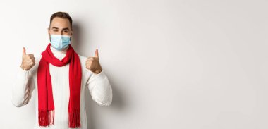 Covid-19, tecrit ve karantina konsepti. Yüz maskesi takan gülümseyen ve baş parmağını kaldıran, kendini koronavirüsten koruyan, beyaz arka planda duran.